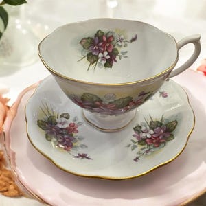 May include: A white teacup and saucer set with a floral pattern of purple and pink flowers. The set has a gold trim around the edges.