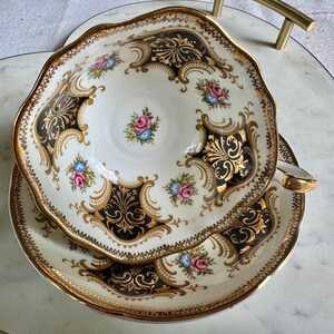 May include: An ornate teacup and saucer set. The white porcelain features black and gold filigree designs, with small floral accents in pink and blue. The edges are scalloped and trimmed with gold.