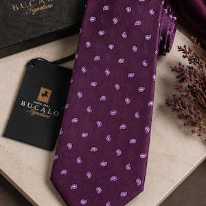 Puede incluir: Una corbata de color morado intenso con un patrón repetitivo de pequeños motivos de cachemira en color morado claro. La corbata viene acompañada de una etiqueta negra de marca y una caja negra a juego, ambas con el texto BUCALO Signature y un logo de caballo.
