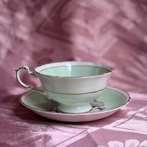 May include: A porcelain teacup and saucer set featuring a pale mint green interior and white exterior with gold trim. The teacup has a scalloped rim and a decorative handle, while the saucer displays a floral pattern in the center.