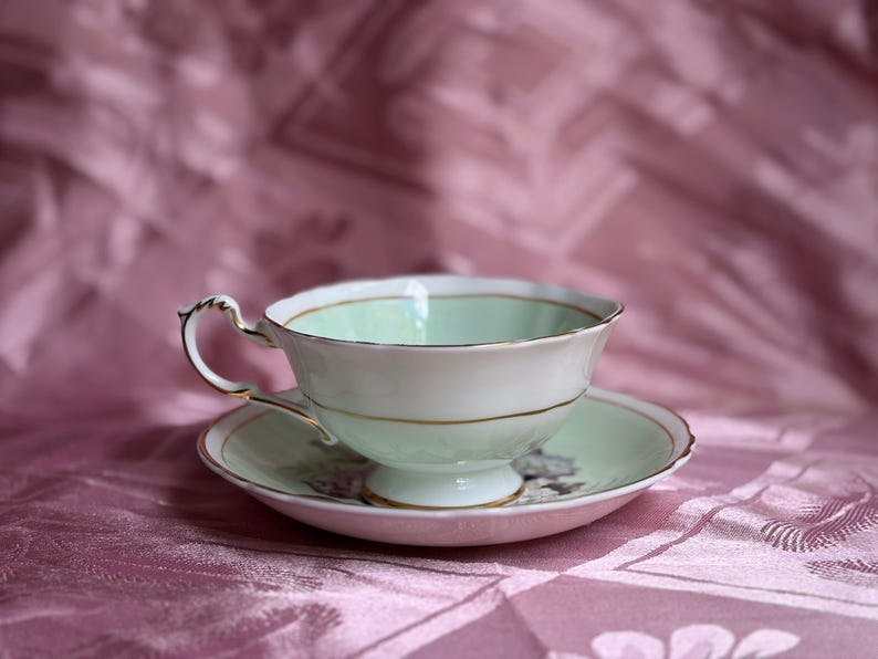 May include: A porcelain teacup and saucer set featuring a pale mint green interior and white exterior with gold trim. The teacup has a scalloped rim and a decorative handle, while the saucer displays a floral pattern in the center.