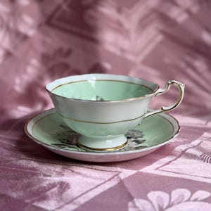 May include: A porcelain teacup and saucer set featuring a pale mint green interior and rim. The exterior of the cup and the saucer display a floral pattern with dark gray details. Both pieces are finished with thin gold trim and a gold handle.