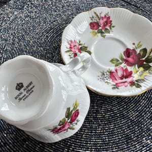 Puede incluir: Un juego de taza y platillo de t&eacute; blanco con un patr&oacute;n floral con rosas rosas y rojas. El platillo tiene un borde festoneado y la taza de t&eacute; tiene un asa. La parte inferior de la taza de t&eacute; tiene las palabras "Royal Albert Bone China England" impresas.