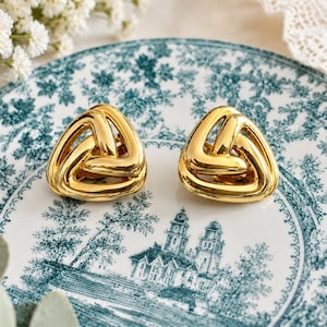 K&ouml;nnte beinhalten: Ein Paar goldfarbene, dreieckige Ohrringe mit geflochtenem Design. Die Ohrringe werden auf einem Teller im Vintage-Stil mit blau-wei&szlig;em Blumenmuster pr&auml;sentiert. Die Ohrringe sind ein auff&auml;lliges Schmuckst&uuml;ck.