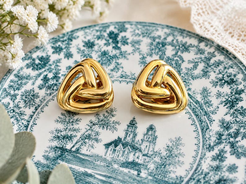 K&ouml;nnte beinhalten: Ein Paar goldfarbene, dreieckige Ohrringe mit geflochtenem Design. Die Ohrringe werden auf einem Teller im Vintage-Stil mit blau-wei&szlig;em Blumenmuster pr&auml;sentiert. Die Ohrringe sind ein auff&auml;lliges Schmuckst&uuml;ck.
