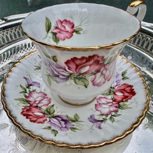 May include: A white teacup and saucer set with a floral design in shades of pink, purple, and green. The rims are trimmed with gold. The set is placed on a silver tray.