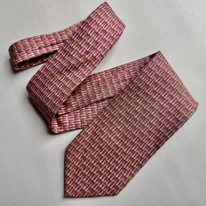 Puede incluir: Una corbata con un patr&oacute;n repetitivo de peque&ntilde;os iconos de anclas de color dorado sobre un fondo morado vibrante. La corbata est&aacute; doblada, mostrando la pala ancha y la parte m&aacute;s estrecha del cuello con un estampado geom&eacute;trico denso.