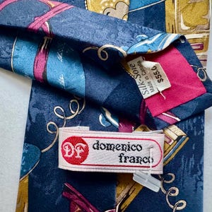 Pu&ograve; includere: Una cravatta blu navy con un motivo astratto multicolore con accenti rosa e oro. La cravatta include un'etichetta in tessuto bianco con il testo Domenico Franco e un logo circolare rosso. Un'etichetta secondaria riporta Silk-Soie Made in Canada.