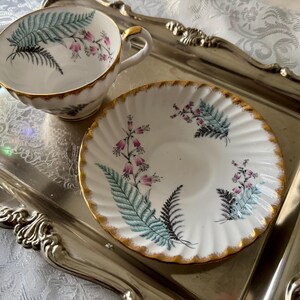 Puede incluir: Juego de taza y platillo de t&eacute; vintage con dise&ntilde;o de helechos y flores. La taza y el platillo de porcelana blanca tienen un borde dorado y est&aacute;n colocados sobre una bandeja plateada. La taza tiene asa y el platillo es redondo.