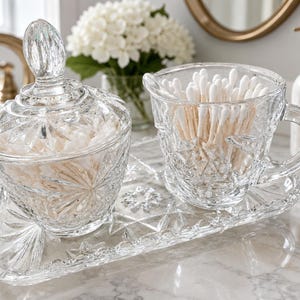 Peut inclure: Un ensemble de toilette en verre transparent composé d'un pot avec couvercle, d'une tasse avec anse et d'un plateau rectangulaire. Chaque pièce présente un motif géométrique et étoilé pressé. Le pot et la tasse contiennent des cotons-tiges.