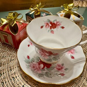 May include: A white teacup and saucer with a floral design featuring pink and red roses. The teacup has a gold rim and the saucer has a scalloped edge. The teacup and saucer are sitting on a gold-colored placemat.