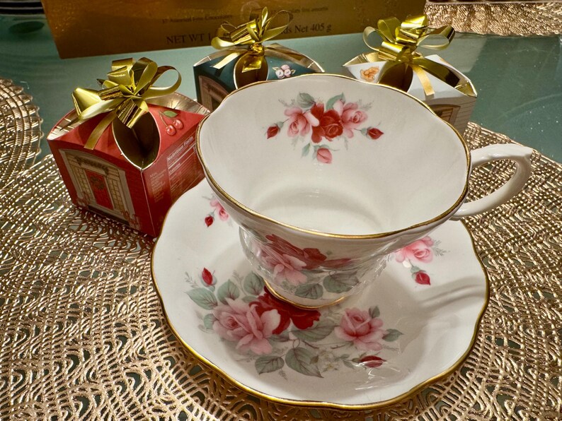 May include: A white teacup and saucer with a floral design featuring pink and red roses. The teacup has a gold rim and the saucer has a scalloped edge. The teacup and saucer are sitting on a gold-colored placemat.