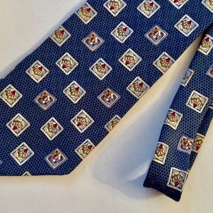 May include: A dark blue necktie featuring a repeating pattern of small, light-colored squares. Each square contains a stylized, fan-shaped motif in shades of gold, white, and red. The background of the tie has a subtle, fine honeycomb texture.