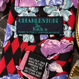 Może przedstawiać: Krawat w czarno-czerwony wz&oacute;r w romby, na kt&oacute;ry nałożono fioletowe i jasnoniebieskie kwiatowe nadruki. Metka zawiera napis Charleston The Rack, Made in Italy, 100% Silk. Dodatkowa metka wskazuje 100% Silk, Made in Italy, Dry Clean Only.