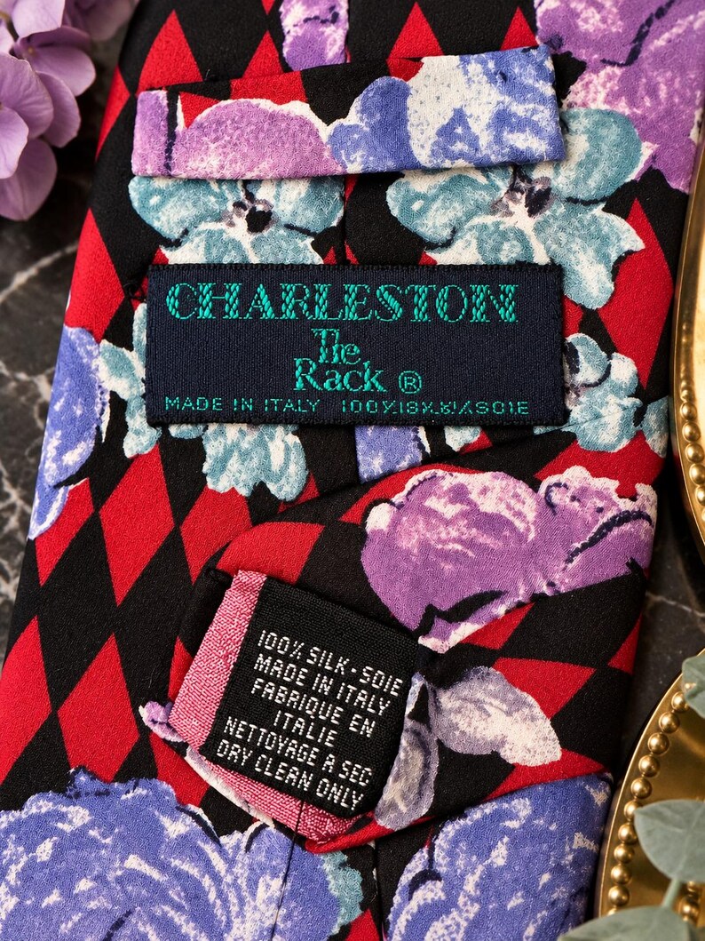 Może przedstawiać: Krawat w czarno-czerwony wz&oacute;r w romby, na kt&oacute;ry nałożono fioletowe i jasnoniebieskie kwiatowe nadruki. Metka zawiera napis Charleston The Rack, Made in Italy, 100% Silk. Dodatkowa metka wskazuje 100% Silk, Made in Italy, Dry Clean Only.