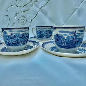 Puede incluir: Tres tazas de t&eacute; y platillos blancos con un dise&ntilde;o de paisaje azul. Las tazas y los platillos tienen un patr&oacute;n azul y blanco detallado alrededor de los bordes y en las superficies. Las tazas de t&eacute; tienen asas curvas.