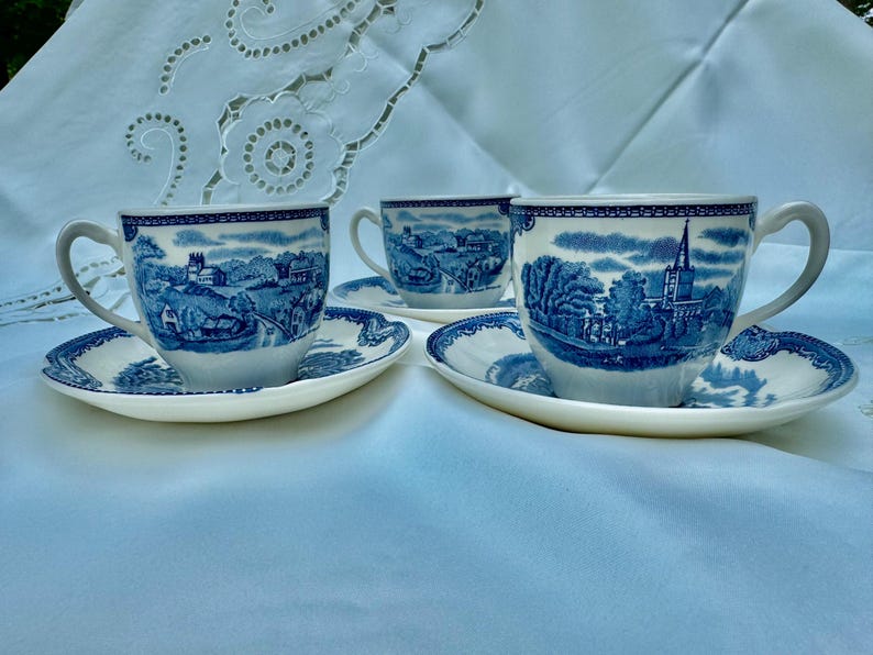 Puede incluir: Tres tazas de t&eacute; y platillos blancos con un dise&ntilde;o de paisaje azul. Las tazas y los platillos tienen un patr&oacute;n azul y blanco detallado alrededor de los bordes y en las superficies. Las tazas de t&eacute; tienen asas curvas.