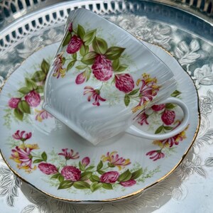 May include: White teacup and saucer set with a floral design featuring pink and yellow flowers and green leaves. The teacup and saucer have gold trim and are set on a silver tray.