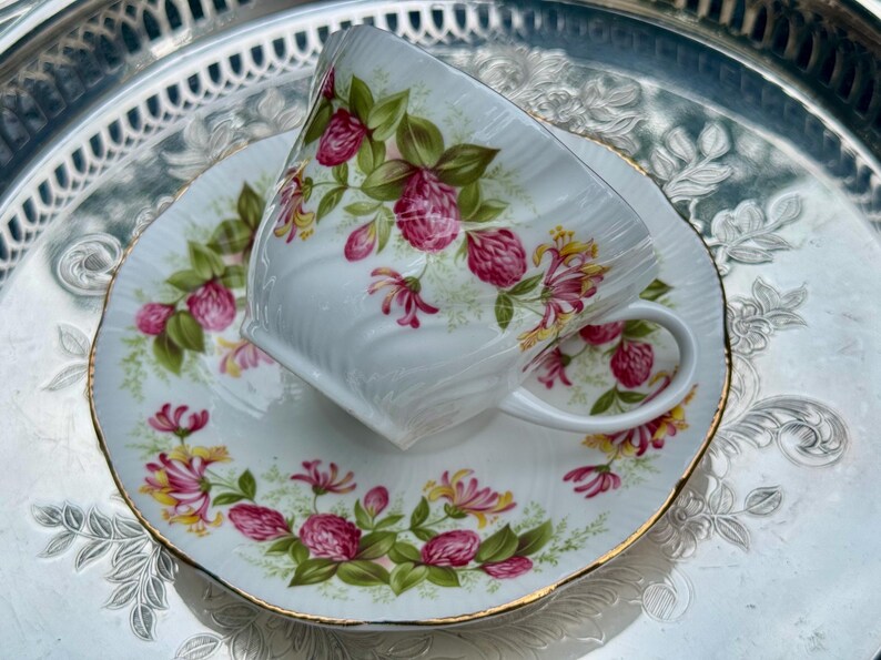 May include: White teacup and saucer set with a floral design featuring pink and yellow flowers and green leaves. The teacup and saucer have gold trim and are set on a silver tray.