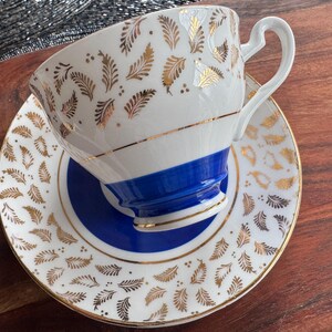 May include: A white teacup and saucer with a blue and gold floral pattern. The cup has a blue band around the top and a gold rim. The saucer has a matching pattern.