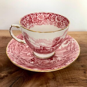 Pu&ograve; includere: Un set vintage di tazza e piattino da t&egrave;. La tazza e il piattino sono bianchi con una scena pastorale rossa e bordi floreali. I bordi sono rifiniti in oro. Il set &egrave; su una superficie di legno.