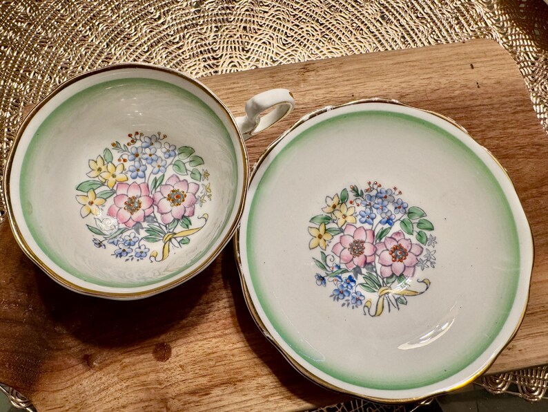 May include: A vintage teacup and saucer set with a green rim and floral design. The floral design features pink, yellow, and blue flowers.