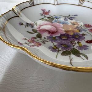 May include: A white, scalloped-edge dish with a gold rim and floral design. The flowers are pink, purple, blue, and yellow. The dish has a delicate, vintage aesthetic.