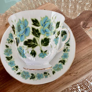 May include: A white teacup and saucer with a floral pattern in shades of blue and green. The floral design features stylised leaves and flowers.