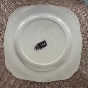 May include: A white, square-shaped plate with a scalloped edge. The underside of the plate is visible, featuring a central recessed area and a black and white label. The plate is set against a textured, light brown surface.
