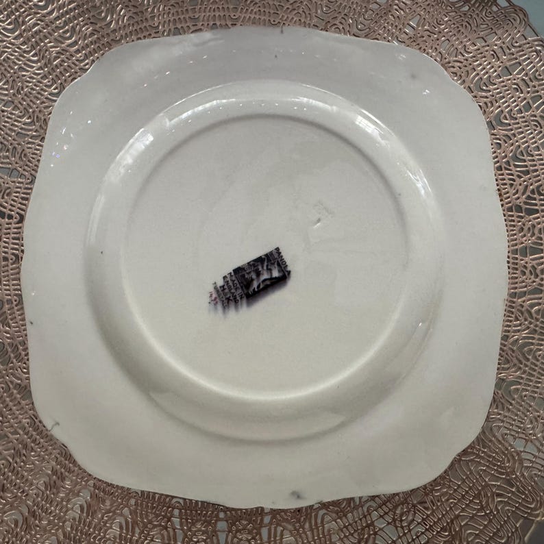 May include: A white, square-shaped plate with a scalloped edge. The underside of the plate is visible, featuring a central recessed area and a black and white label. The plate is set against a textured, light brown surface.