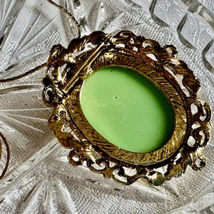 May include: A vintage gold-toned brooch with an oval, light green center. The brooch has an ornate, filigree-style frame. A delicate gold chain is attached, suggesting it can be worn as a pendant. The brooch is set against a textured glass surface.