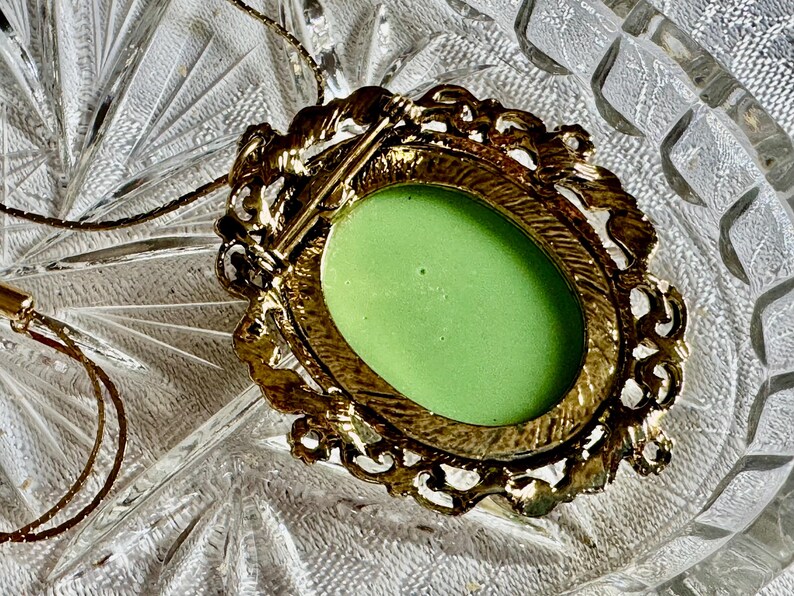 May include: A vintage gold-toned brooch with an oval, light green center. The brooch has an ornate, filigree-style frame. A delicate gold chain is attached, suggesting it can be worn as a pendant. The brooch is set against a textured glass surface.