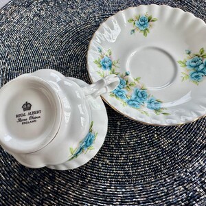May include: A white teacup and saucer with a blue floral pattern. The teacup has a scalloped rim and the saucer has a gold trim. The bottom of the teacup has the words "Royal Albert Bone China England" printed on it.