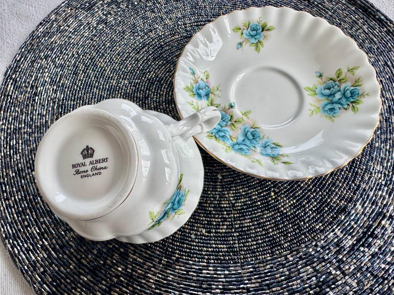 May include: A white teacup and saucer with a blue floral pattern. The teacup has a scalloped rim and the saucer has a gold trim. The bottom of the teacup has the words "Royal Albert Bone China England" printed on it.