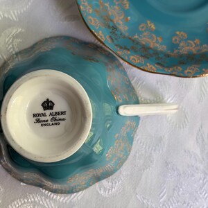 Może przedstawiać: Turkusowy zestaw filiżanek ze spodkiem z porcelany kostnej ze złotym wzorem kwiatowym. Sp&oacute;d filiżanki zawiera czarny napis ROYAL ALBERT BONE CHINA ENGLAND z ikoną korony. Filiżanka posiada białe ucho oraz falowane krawędzie.