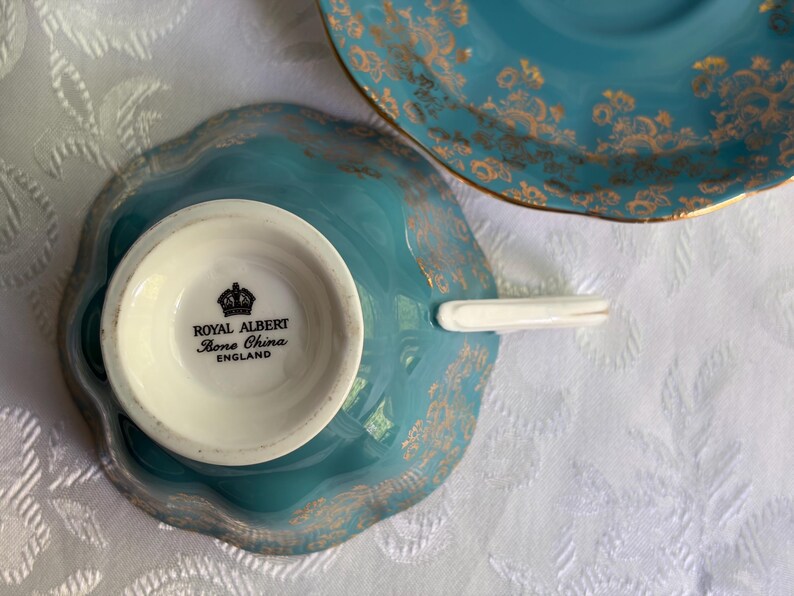 Może przedstawiać: Turkusowy zestaw filiżanek ze spodkiem z porcelany kostnej ze złotym wzorem kwiatowym. Sp&oacute;d filiżanki zawiera czarny napis ROYAL ALBERT BONE CHINA ENGLAND z ikoną korony. Filiżanka posiada białe ucho oraz falowane krawędzie.
