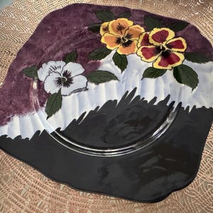 May include: Square decorative plate with a painted floral design. The plate features three pansies in shades of yellow, burgundy, and white, with green leaves. The background has a dark purple, white, and black color scheme.