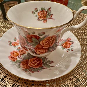 Pode incluir: Uma ch&aacute;vena de ch&aacute; e um pires brancos com uma borda dourada e um padr&atilde;o floral. O padr&atilde;o floral apresenta rosas cor-de-rosa e vermelhas com folhas verdes.