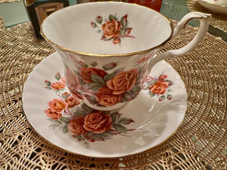 Pode incluir: Uma ch&aacute;vena de ch&aacute; e um pires brancos com uma borda dourada e um padr&atilde;o floral. O padr&atilde;o floral apresenta rosas cor-de-rosa e vermelhas com folhas verdes.