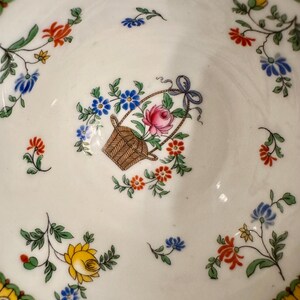 Pu&ograve; includere: Una tazza da t&egrave; in porcellana bianca con un motivo floreale. Il motivo presenta un cesto di fiori con una rosa rosa, fiori blu e foglie verdi. La tazza &egrave; rifinita con un bordo giallo e verde.