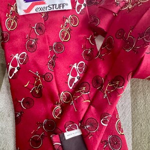 May include: A bright red necktie featuring a repeating pattern of black and yellow bicycles. The tie includes a white label with the text "exerSTUFF" and a smaller white tag near the bottom edge that reads "MADE IN CHINA" in black capital letters.