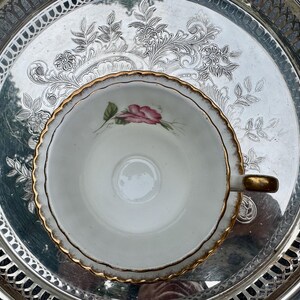 May include: A vintage teacup with a pink rose design and gold trim sits on a silver serving tray. The tray has an ornate floral pattern around the edge and center. The teacup handle is gold.