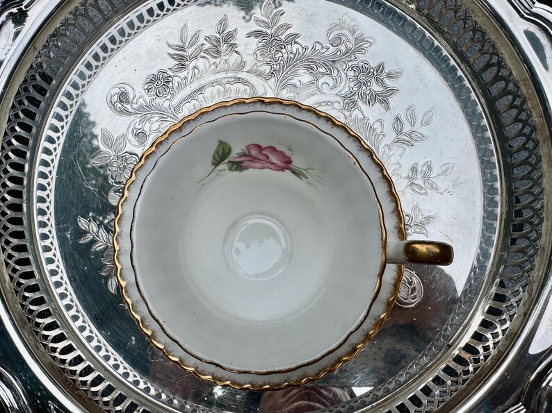 May include: A vintage teacup with a pink rose design and gold trim sits on a silver serving tray. The tray has an ornate floral pattern around the edge and center. The teacup handle is gold.
