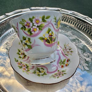 May include: A white teacup and saucer set with floral and ribbon designs, resting on a silver tray. The teacup and saucer feature pink and yellow flowers with green leaves and pink ribbons. The teacup has a gold rim.