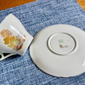 May include: A white porcelain teacup and saucer set featuring a colorful landscape illustration of trees and foliage on the cup. The saucer is turned upside down, revealing a green Shelley England maker mark and the word Heather printed on the base.