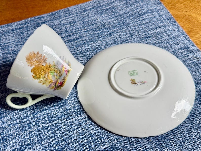 May include: A white porcelain teacup and saucer set featuring a colorful landscape illustration of trees and foliage on the cup. The saucer is turned upside down, revealing a green Shelley England maker mark and the word Heather printed on the base.