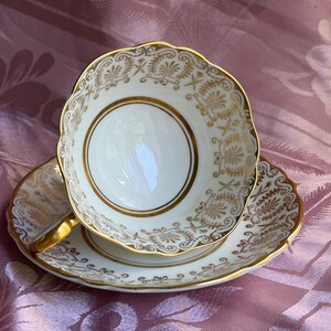 May include: A white porcelain teacup and saucer set featuring a scalloped edge and an intricate gold filigree pattern. The cup has a gold-trimmed handle and a circular gold band inside the base, with matching gold decorative motifs on the saucer.