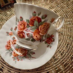 Pode incluir: Uma ch&aacute;vena de ch&aacute; e um pires brancos com um padr&atilde;o floral de rosas laranja e rosa. A ch&aacute;vena de ch&aacute; tem uma borda dourada e uma pega delicada.