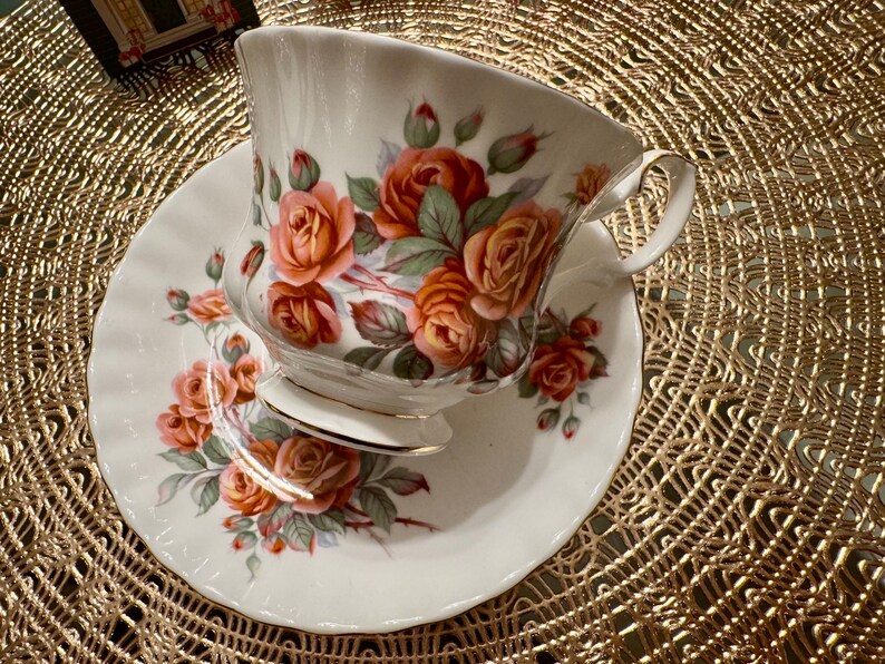 Pode incluir: Uma ch&aacute;vena de ch&aacute; e um pires brancos com um padr&atilde;o floral de rosas laranja e rosa. A ch&aacute;vena de ch&aacute; tem uma borda dourada e uma pega delicada.