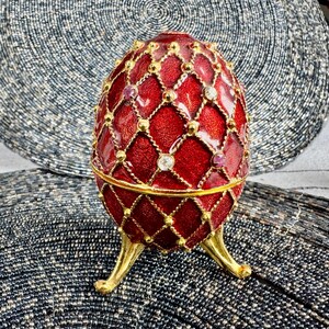 May include: A red and gold jeweled egg with a decorative diamond pattern. The egg is on a stand with three gold legs.